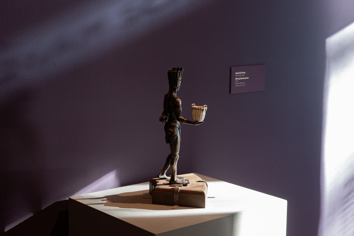 A small bronze sculpture of a standing Indigenous figure wearing a feathered headdress is positioned on a wooden base atop a white pedestal. The figure is shown in profile, extending one hand forward to hold a small, delicately woven light-colored basket. Soft, directional light from the right casts dramatic shadows across the wall and the sculpture, highlighting the fine detail of the bronze work and the texture of the miniature basket.