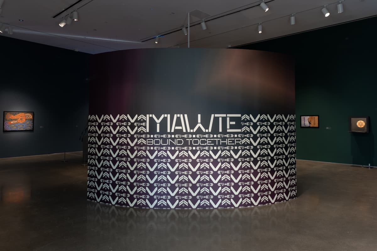 A large, cylindrical wall in a gallery featuring a dark purple and green gradient background covered in intricate white geometric patterns. Bold, stylized white text in the center reads "MAWTE BOUND TOGETHER". The surrounding space displays several other artworks on dark green walls, including a framed piece and an open shadow box containing a circular pattern.