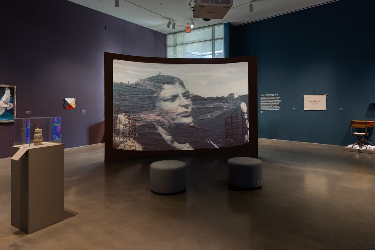 A projection on a large, curved wall displays a black-and-white portrait of an Indigenous woman's face, partially overlaid with moving water ripples and geometric patterns. In front of the screen, two grey cylindrical ottomans provide seating on a polished concrete floor, while the surrounding gallery walls in shades of plum and deep teal feature various sculptures and wall-mounted artworks. To the left, a small woven basket is displayed in a clear acrylic case atop a tall grey pedestal.