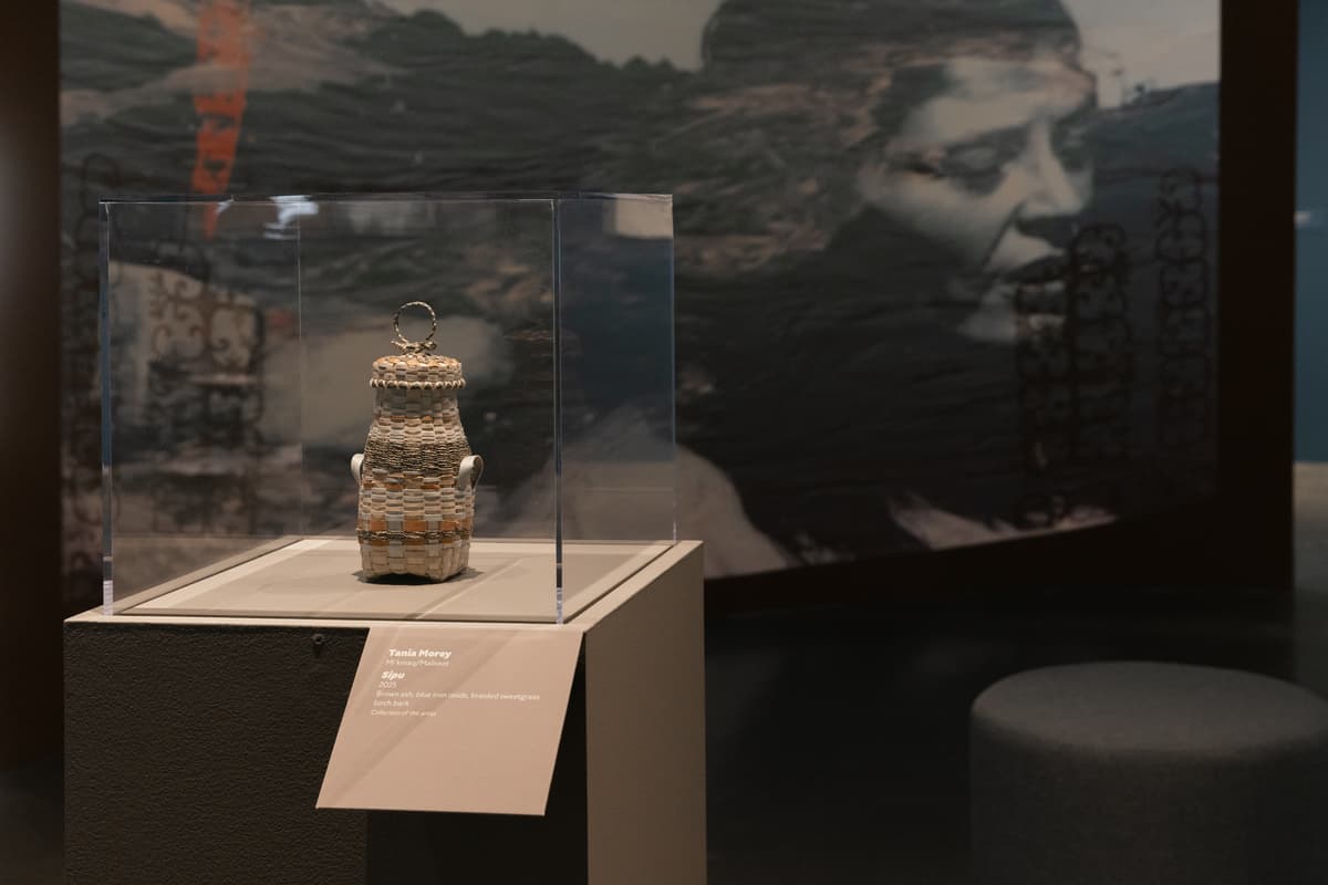 A small, intricate basket titled displayed within a clear acrylic case on a grey pedestal. The woven vessel, crafted from brown ash, blue iron oxide, braided sweetgrass, and birch bark, features subtle color bands and delicate side handles. In the background, a large curved screen displays a blurred black-and-white portrait of a woman, while a grey ottoman sits on the polished gallery floor.