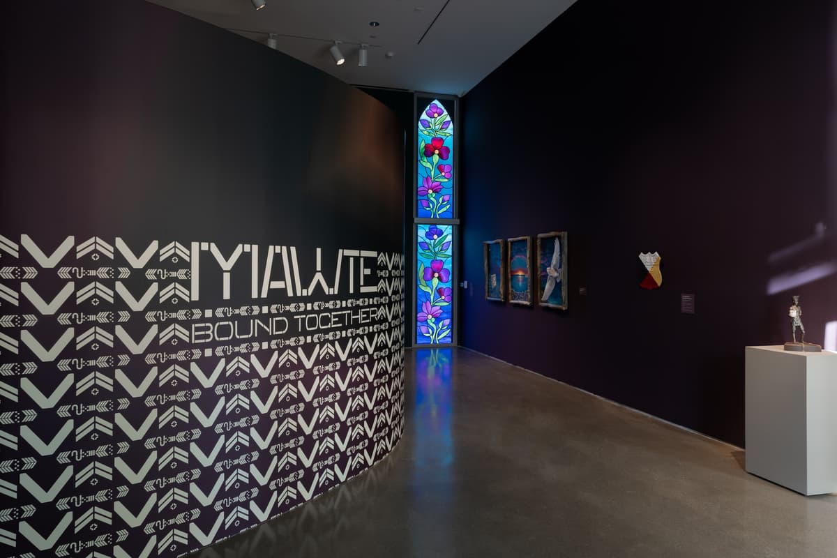A dark, curved installation wall with a black-and-white graphic pattern of repeating stylized symbols. In the center, the bold white text reads "MAWTE BOUND TOGETHER". To the right, three vertical paintings in rustic wooden frames are followed by a painted shield-shaped sign and a bronze sculpture on a white pedestal. The composition is framed by a tall stained-glass window at the far end of the hallway that features vibrant purple flowers and reflects light onto the polished floor.