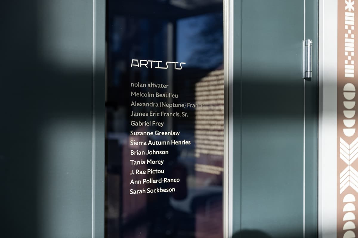 A glass gallery door with the word "ARTISTS" in a stylized, outlined white font, followed by a list of names in solid white text: nolan altvater, Melcolm Beaulieu, Alexandra {Neptune} Francis, James Eric Francis, Sr., Gabriel Frey, Suzanne Greenlaw, Sierra Autumn Henries, Brian Johnson, Tania Morey, J. Rae Pictou, Ann Pollard-Ranco, Sarah Sockbeson