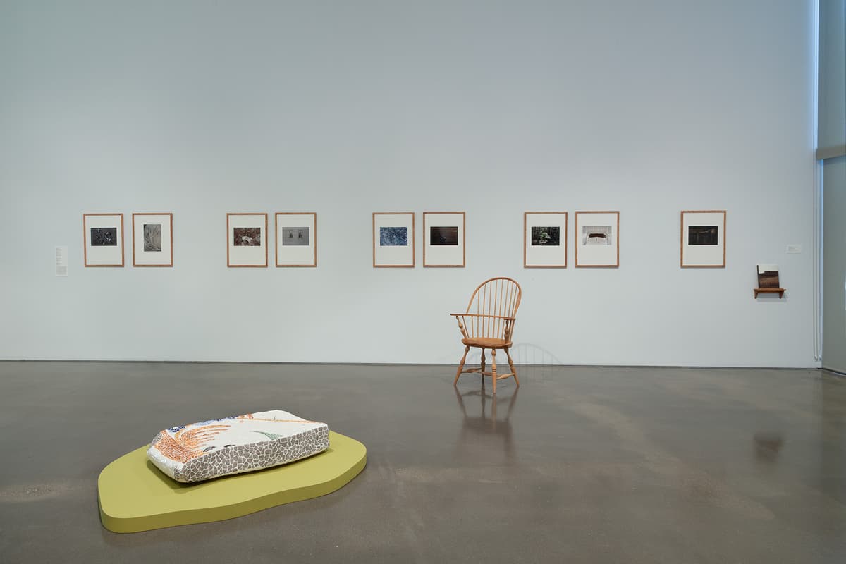 An installation view of a well-lit art gallery with white walls and a light wood floor. On the main wall, three large paintings are displayed side by side: a vertical landscape of bare winter tree branches, a vertical painting of a tree trunk surrounded by bright green leaves, and a horizontal painting of a calm blue sea against a beige shoreline. In the center of the room sits a long black tufted leather bench under bright track lighting.