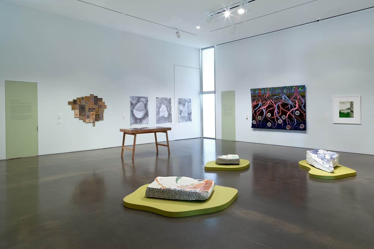 An installation view of a gallery corner featuring a white wall meeting a bright coral-pink wall over light hardwood flooring. The white wall displays two large horizontal landscape drawings in simple wooden frames. The coral-pink wall on the right showcases a wide horizontal sequence of smaller framed sketches and colorful paintings.