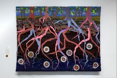 Installation view of a large-scale, textured textile piece mounted on a wall. The artwork portrays a landscape split between the surface world and the underground. At the top, slender trees with green leaves stand along a rocky stone wall or shoreline against a blue sky. Below the stones, a sprawling and thick root system rendered in bright pink, lavender, and sky blue yarn extends deeply into a dark brown and deep blue subterranean layer. Scattered among the roots are approximately eight small, circular, multi-colored embroidered medallions.