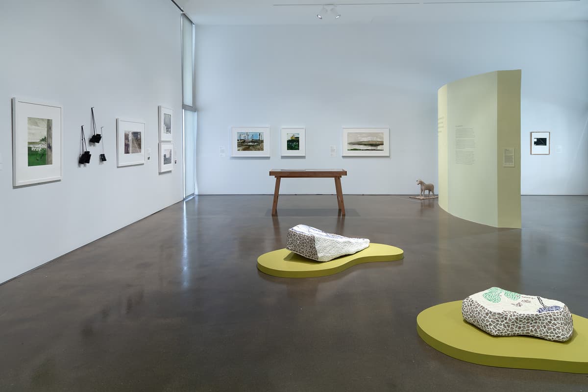 An installation view of a coral-pink gallery wall showing a large, rectangular grid layout of eighteen small white-framed minimalist sketches under track lighting. The artwork arrangement hangs uniformly above a light-colored hardwood floor. A wooden bench sits obliquely in the foreground.