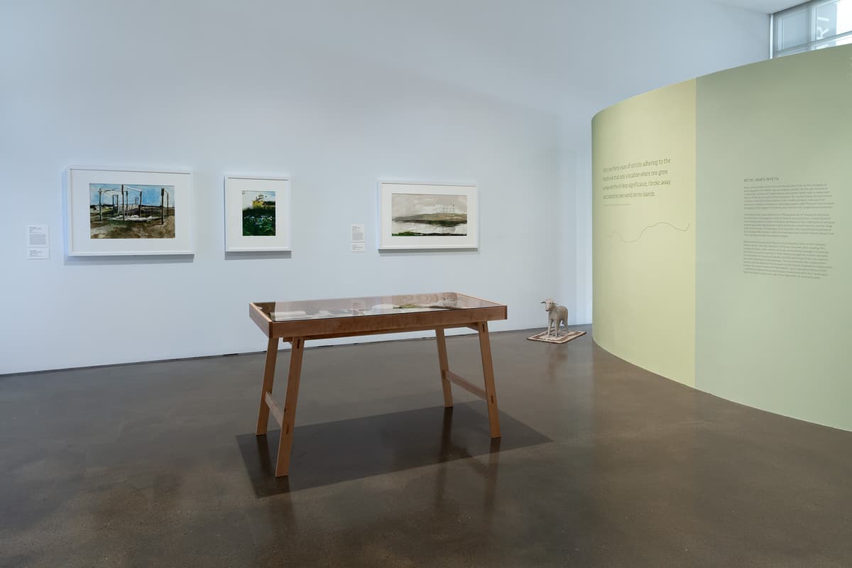 An installation view of a corner where a coral-pink wall meets an off-white wall over a light hardwood floor. The pink wall on the left displays a large vertical grid of eighteen small white-framed sketches, while the off-white wall on the right features two larger framed sketches. A long wooden gallery bench sits positioned obliquely in the room under overhead track lighting.