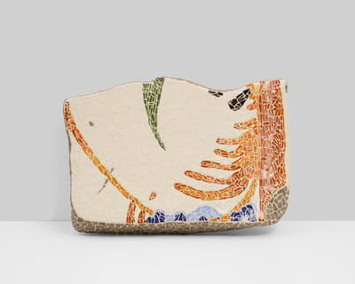 Irregular horizontal ceramic mosaic with an off-white background. Abstract designs in orange, green, and blue tesserae are scattered across the surface, which is set into a rough, sandy-brown base.