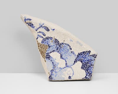 A free-standing, sculptural mosaic fragment with an irregular, trapezoidal shape that tapers to a point at the upper left. The piece is composed of "pique assiette" style ceramic shards. The primary surface is covered in white matte tiles, with a flowing, organic pattern of blue and light blue glazed ceramic pieces moving across the center. A small section of textured, metallic gold or bronze-toned shards is visible on the left edge. The object is set against a neutral, two-toned grey and white background.