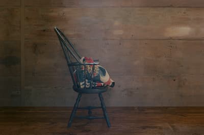 A profile view of a dark green or black Windsor chair centered against a wall of wide, horizontal wooden planks. A patterned textile with red and cream tones is draped over the seat and armrest. The wooden wall in the background shows faint scrawled markings and inscriptions. The scene is set on a dark, rustic wooden floor.