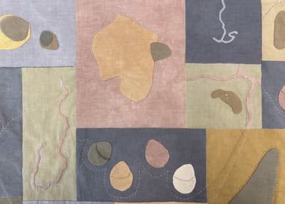 A close-up detail of a textile work featuring a patchwork of rectangular linen panels in muted tones of lavender, dusty rose, sage green, slate grey, and mustard yellow. Various organic, pebble-like shapes in contrasting earth tones are appliquéd onto the panels using visible stitching. Delicate, wandering lines of white dashed embroidery and pink fabric piping travel across the composition, suggesting cartographic features like paths or coastlines. The texture of the woven fabric is prominent throughout.