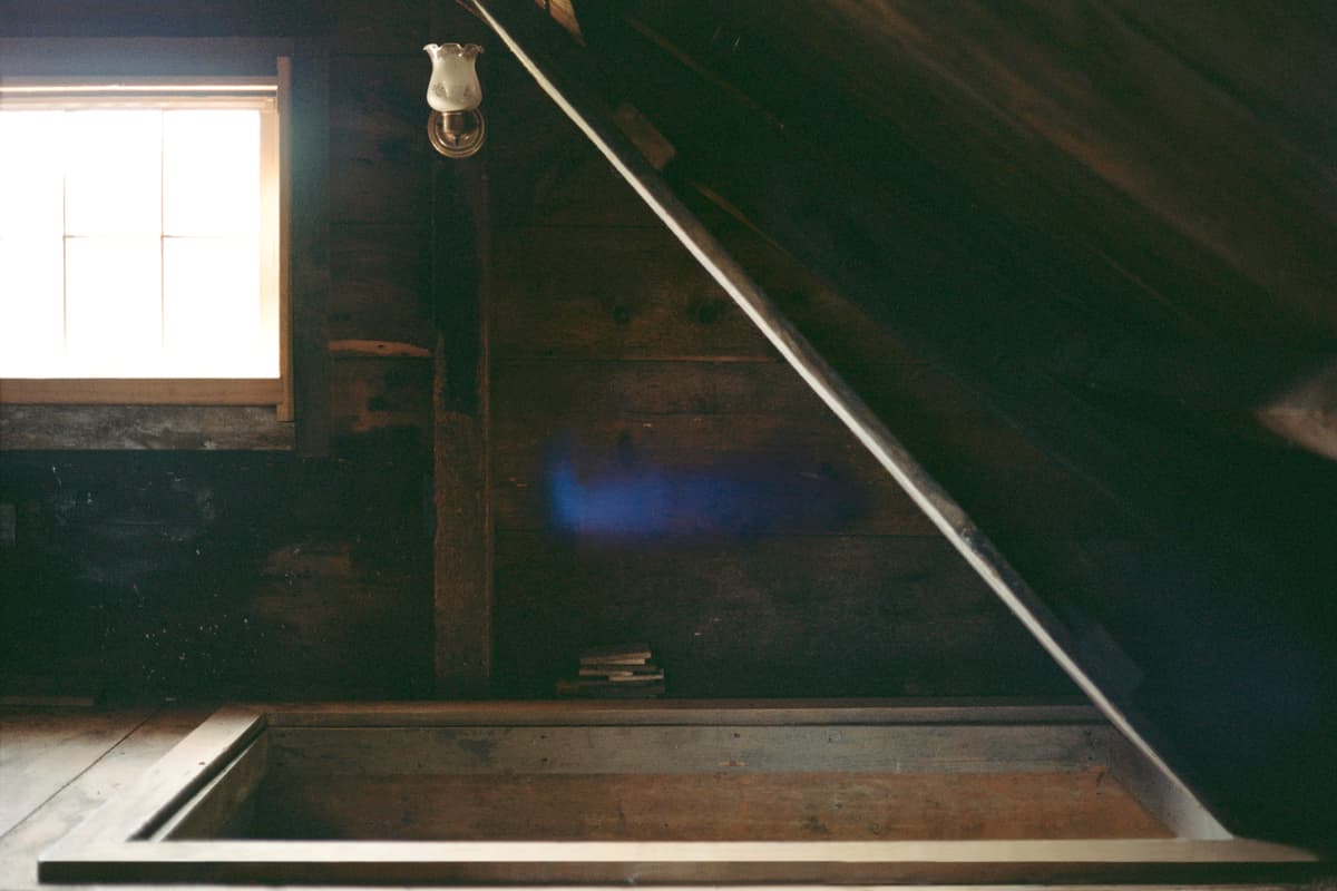 A photograph of a dimly lit, rustic wooden attic space. In the center of the dark wooden floor, an open rectangular hatch leads to the floor below. A small window on the left lets in bright white light, and a single lit wall sconce glows near a slanted ceiling beam. A mysterious, faint blue horizontal blur floats in the center of the frame.