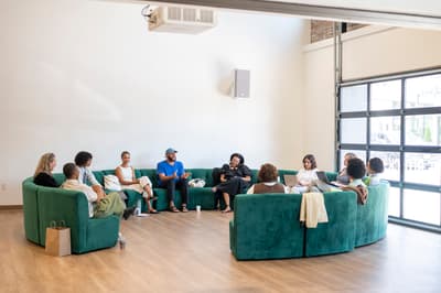 A group of about ten people are seated in a large circle on a curved, emerald green velvet sectional sofa. They are gathered in a bright, minimalist room with light wood floors and high white walls. A large glass garage-style door on the right lets in natural light, and a projector is mounted on the ceiling. The atmosphere appears casual and collaborative; some individuals are talking and gesturing, while others listen or look at laptops. The group is dressed in relaxed, everyday clothing. A brown paper gift bag sits on the floor in the foreground.
