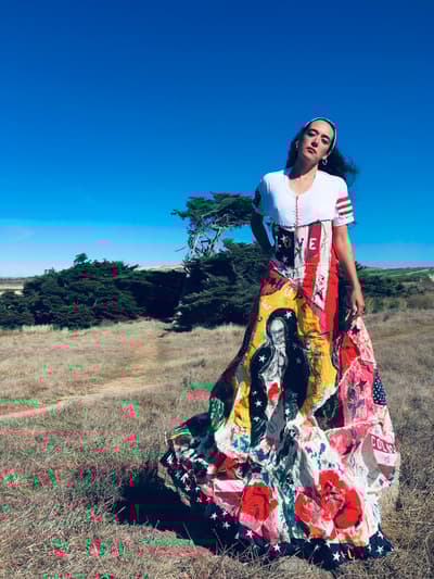 A woman stands in a dry, grassy field under a bright blue sky, wearing a long, voluminous skirt made from a collage of colorful fabrics. The skirt features bold graphics including the word "LOVE," American flag motifs, yellow sections, and a large black and white illustration of a figure resembling the Virgin Mary. She is also wearing a white t-shirt with red and white striped sleeves. In the background, there are wind-sculpted evergreen trees and rolling hills under a clear, cloudless sky.