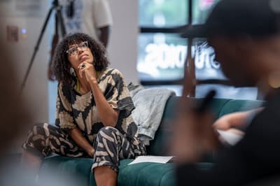 A woman with dark, curly hair and clear glasses sits on a green velvet sofa, looking upward and to the right with her hand to her chin in a contemplative pose. In the background, there is a large window and a professional camera setup on a tripod is partially visible.
