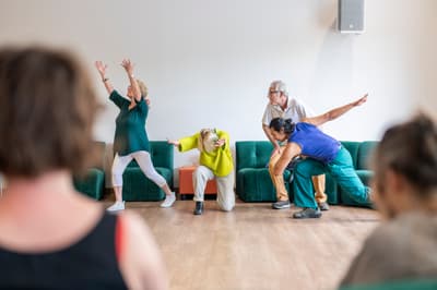 Four people of various ages perform expressive movements in a bright studio space. In the center, a person in a bright yellow sweater kneels with arms outstretched, while others around them strike dynamic poses with reaching arms. The audience is visible as blurred figures in the foreground, and the signature teal velvet seating lines the back wall.