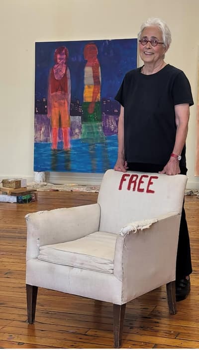 Artist Katherine Bradford stands in her studio, smiling and resting her hands on the back of a worn, white upholstered armchair. The word "FREE" is painted in red across the top of the chair's backrest. In the background, one of her large-scale paintings is visible, depicting two abstracted figures standing in water against a dark blue night sky. The studio features original hardwood floors and a white wall, capturing a bright, creative environment.