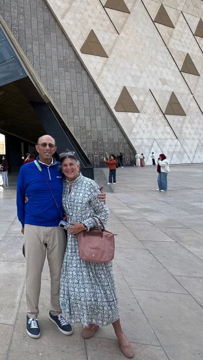 A full-length photo of Barbara and Ted Alfond smiling together in an outdoor plaza. Ted, on the left, wears a bright blue quarter-zip pullover, tan trousers, and dark sneakers. Barbara, on the right, wears a long-sleeved, patterned dress in white and teal, carrying a pink handbag. They stand before the Grand Egyptian Museum, which features a massive, modern sloped wall behind them decorated with hieroglyphics and triangular patterns. Other visitors are visible in the background of the wide, stone-paved courtyard.