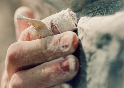 Close-up view of a hand holding an object, featuring taped knuckles and fingertips with white and red chalk. The tape is layered over the red skin of the middle and index fingers, showing texture and dust. The object is a white, fibrous mass, potentially cotton or gauze, next to a rough, darker surface.