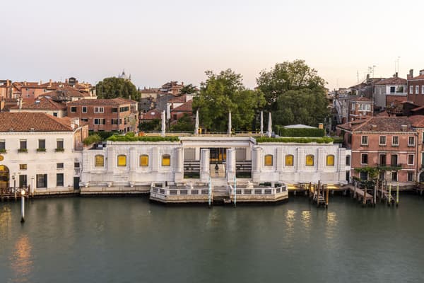 A horizontal, single-story white Istrian stone building on a waterfront, characterized by a repetitive rhythm of arched windows and a central recessed entrance flanked by four columns. A roof terrace with a green hedge and white umbrellas sits atop the structure, which is nestled among taller terracotta-roofed buildings.