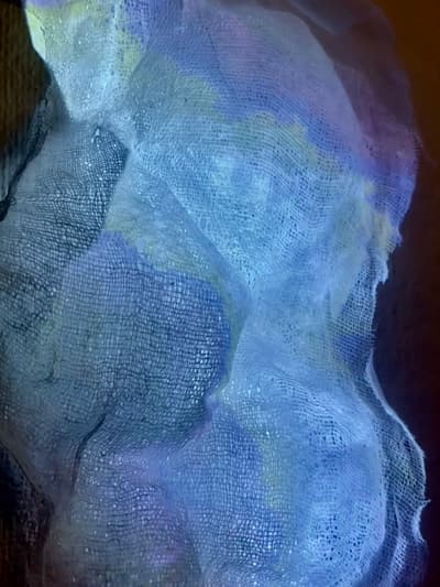 Close-up photograph of a layered and folded piece of dyed cheesecloth or gauze. The fabric features varying blue, purple, and green pigments. The weave is loose and open, with frayed edges, displayed against a dark, contrasting background.