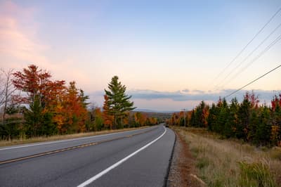 Landscape photograph from the perspective of a two-lane paved road. The road has yellow center lines and white shoulder lines, curving toward the right. It is flanked by a gravel shoulder and a mix of deciduous trees with red and orange autumn foliage on the left, and coniferous trees on the right. Power lines and wooden poles run along the right side under a pale sky.