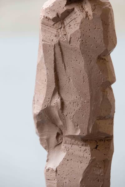 Frontal view of a hand-sculpted clay form, with numerous irregular facets and tool marks. The surface is raw and textured with visible pinholes and grooves, tapering towards a base. The object is centered against a solid light blue-grey background.