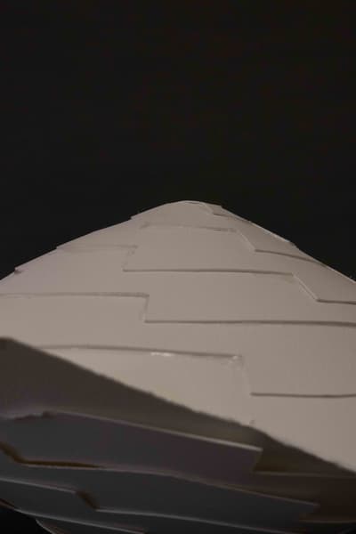 Photograph of a white paper or card prototype, featuring a series of overlapping step-like layers that form a central spine. The structure extends from a blurred foreground towards a sharp, pointed top, against a dark black background. The cuts and layers suggest a model for architectural or folded design.