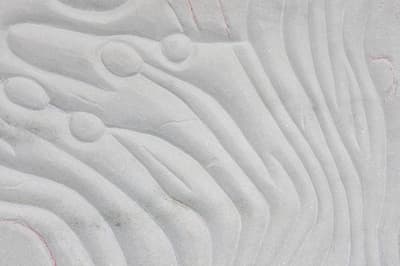 Macro photograph of a light grey stone or plaster surface. The material is carved with deep, rhythmic undulating ridges that mimic water ripples or organic folds. Several small, raised circular nodules are integrated into the textured lines, with faint red pigment visible within the recessed grooves.