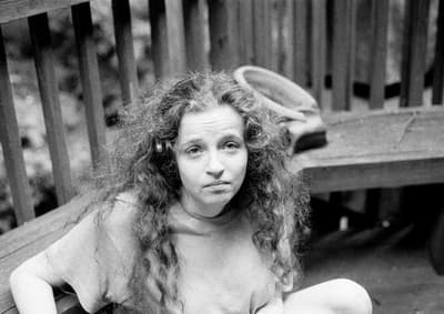 Black and white candid portrait of a young woman with voluminous, curly hair sitting on a wooden deck. She is wearing a light-colored t-shirt and looking directly at the camera with a neutral expression. The background features vertical wooden slats of a deck railing, slightly out of focus.