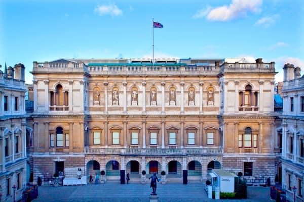 A symmetrical, three-story Neo-Palladian stone building with a rusticated ground floor supporting a piano nobile with Corinthian pilasters and arched niches containing figurative sculptures. A central flagpole with a British flag tops the balustraded roofline, overlooking a paved courtyard.