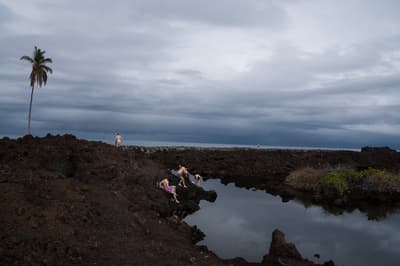 A landscape photograph of a dark, jagged volcanic rock formation bordering a calm pool of water. A group of four people in swimwear are positioned on the rocks near the water's edge. A single, tall palm tree stands on the left horizon under an expansive, heavy grey sky with dense cloud cover.