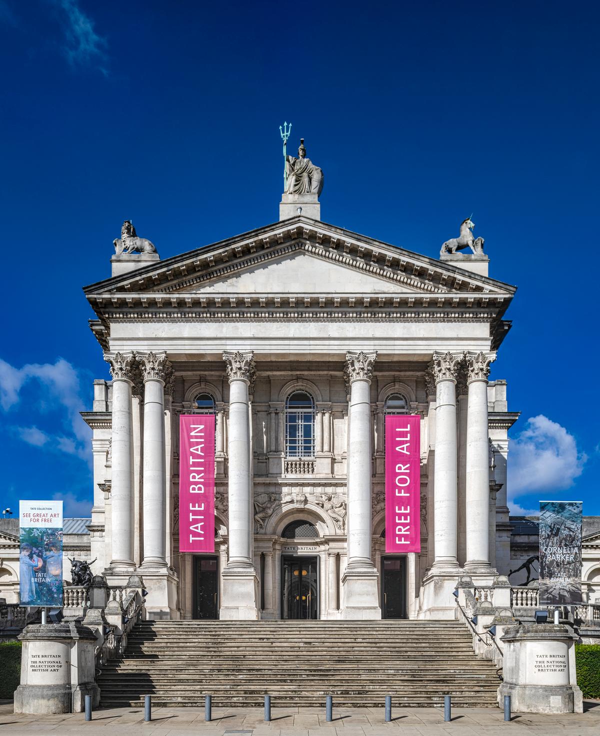 A grand Neoclassical facade featuring a deep portico with eight Corinthian columns supporting a triangular pediment. A central dome is topped with a statue of Britannia, and a wide flight of stone stairs leads to the entrance. Two vertical pink banners hang between the columns.
