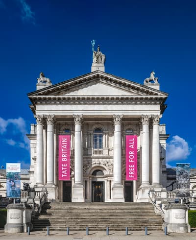 A grand Neoclassical facade featuring a deep portico with eight Corinthian columns supporting a triangular pediment. A central dome is topped with a statue of Britannia, and a wide flight of stone stairs leads to the entrance. Two vertical pink banners hang between the columns.