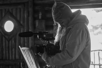 Black and white photograph of a person in a profile view, wearing a knit beanie and a heavy jacket. They are looking down while holding a professional cinema camera equipped with a large shotgun microphone. The scene is indoors with a rustic, log-cabin style wall and a bright, overexposed window in the background providing strong rim lighting.
