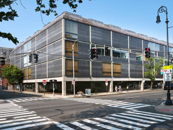 A four-story rectangular building featuring a modular grid of matte pewter-finished stainless steel panels and reflective glass windows. The concrete frame is exposed at the ground level to create an open pedestrian colonnade at a city street corner.