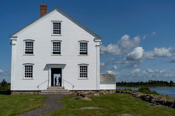 A three-story, white clapboard historic house stands on a grassy lawn next to a body of water under a blue sky with fluffy clouds. A gravel path leads to the open front door, through which the silhouettes of two people can be seen looking out the back of the house toward the distant, tree-lined shore.