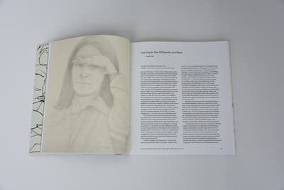 An open book features a full-page graphite portrait on the left and text on the right. The left page shows a soft, detailed drawing of a woman (Ada) shielding her eyes with her hand, looking directly at the viewer. The right page contains an essay titled "Learning to See Differently and More" by Kiko Aebi, set in a clean serif typeface with significant white space around the margins.