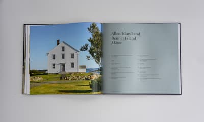 A landscape-oriented open book rests on a white surface. The left page features a full-bleed color photograph of a three-story white clapboard house on Allen Island, Maine, under a clear blue sky. A stone wall and grassy path lead to the centered front door. The right page is a pale blue-grey, featuring the title "Allen Island and Benner Island, Maine" in a black serif font, followed by a two-column list of essay titles and authors.
