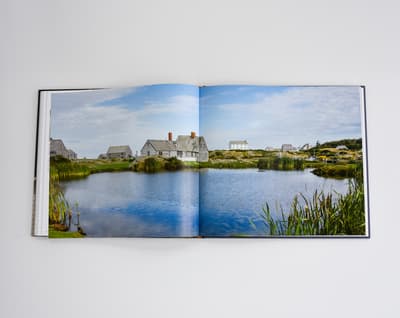 An open book shows a panoramic color photograph spanning both pages. The image depicts a cluster of weathered, grey wooden houses and outbuildings situated on a rocky shoreline in Maine. In the foreground, a calm body of water reflects the blue sky and white clouds. Tall green reeds grow along the water’s edge in the lower right and left corners.