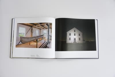 An open book displays two contrasting photographs. The left page shows the interior of a rustic wooden building with exposed ceiling beams and light wood floors. A large, historic birchbark canoe is displayed on a wooden trestle in the center of the room. The right page features a full-bleed photograph of a white house at night, illuminated by a bright light source from the front against a pitch-black, starry sky.