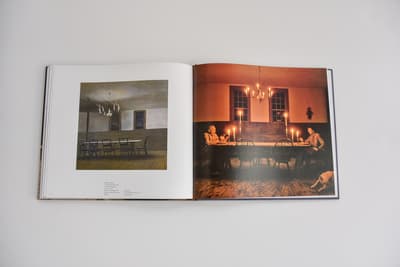 An open book featuring two interior scenes centered around long dining tables. The left page shows a painting by Andrew Wyeth, The Evening Meal, depicting a spare, dimly lit room with a long table and a chandelier. The right page features a full-bleed color photograph of a similar room, warmly lit by many candles on a long table where two people sit at opposite ends, creating a glow against the dark wooden walls.