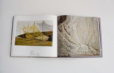 An open book showing two images related to maritime textures. The left page is a painting of yellow-toned fishing nets stretched out to dry on tall poles along a coastline. The right page is a full-bleed, close-up photograph of white, intricately knotted netting bunched together, highlighting the geometric patterns and shadows of the mesh.