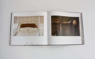 An open book with two photographs. The left page shows a bed with white linens and a decorative lace or fringed canopy hanging above the headboard. The right page is a dark, moody photograph of a wooden interior wall where a woven basket and pieces of driftwood are mounted as decor.