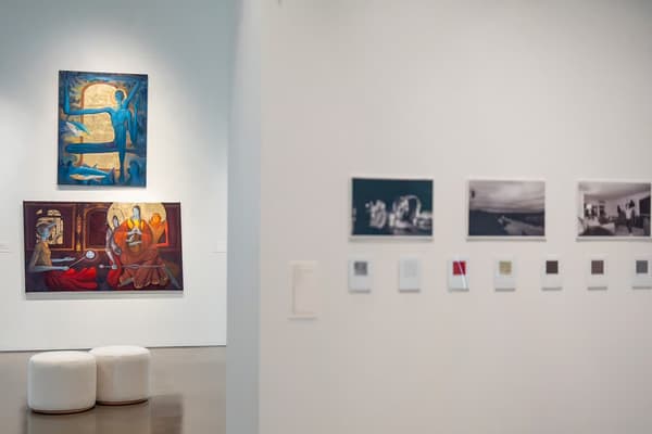An exhibition view inside a modern art gallery, split by a foreground wall. On the left wall in the background, two large, colorful paintings featuring stylized figures and gold leaf are stacked vertically above two small, white cylindrical stools. On the right foreground wall, a row of black-and-white photographs hangs above a series of small, square prints.