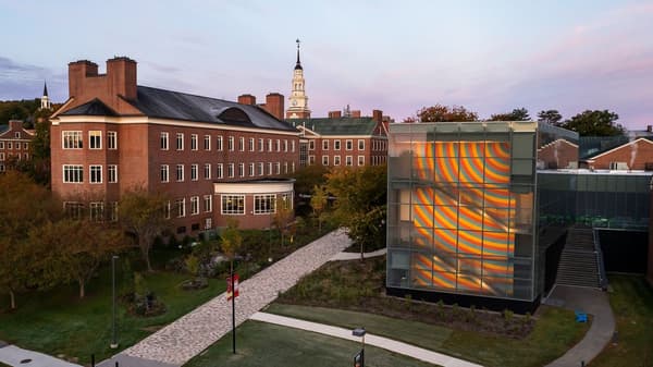 An elevated view of a Colby campus at dusk featuring traditional red brick buildings and a modern glass structure. Inside the glass building, a large-scale mural with colorful, curved stripes of yellow, orange, and blue is visible across multiple floors. A cobblestone path leads through a landscaped lawn with trees, set against a soft purple and pink evening sky with a clock tower in the distance.