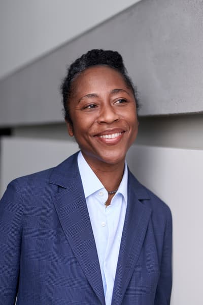 A professional headshot of Naomi Beckwith. She is smiling and looking off-camera to the right. She has short, dark hair and is wearing a blue patterned blazer over a light blue button-down shirt. The background consists of minimalist, neutral-toned architectural panels.