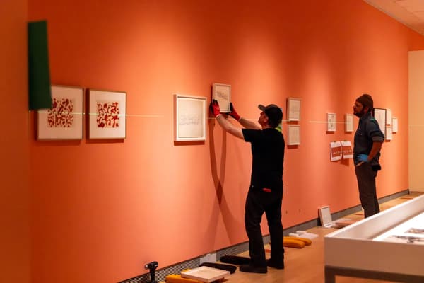 An eye-level, wide shot captures two art preparators installing an exhibition inside a gallery with vibrant peach-colored walls.
