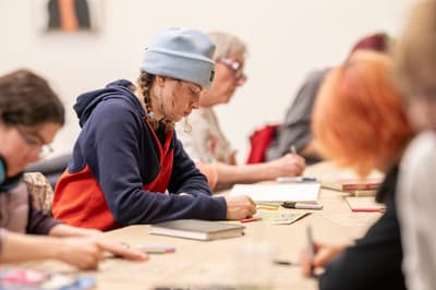 A medium, eye-level shot of several people drawing at a long wooden table. The central figure has braided hair, a light blue beanie, and a navy and red hoodie, looking down at a sketch. Other figures are seated along the table, appearing blurred in the foreground and background. Sketchbooks, markers, and colored pencils are scattered across the table surface.