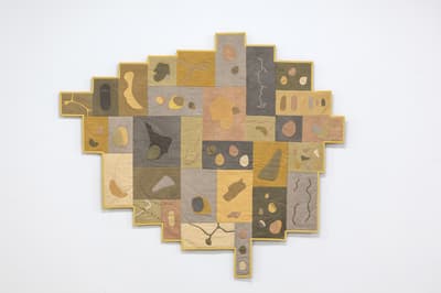 A textile work featuring a patchwork of rectangular linen panels in muted tones of lavender, dusty rose, sage green, slate grey, and mustard yellow. Various organic, pebble-like shapes in contrasting earth tones are appliquéd onto the panels using visible stitching. Delicate, wandering lines of white dashed embroidery and pink fabric piping travel across the composition, suggesting cartographic features like paths or coastlines. The texture of the woven fabric is prominent throughout.